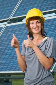 Upper body shot of a young brunette woman in a short-sleeved casual dress and yellow construction helmet in front of a free-standing photovoltaic system showing both thumbs up