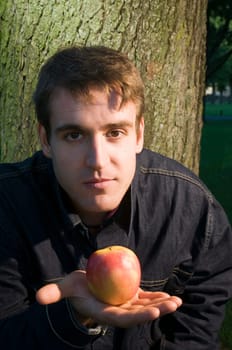 Head and shoulders shot of a young man leaning against a tree towards the camera,offering an apple