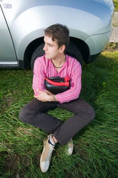 Young man sitting at the front wheel of a vehicle with a black gasoline canister and looking to the side helplessly