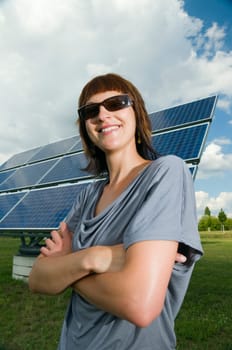 Wide angle torso shot of a young brunette woman in casual casual dress with crossed arms in front of photovoltaic system on green meadow and cloudy sky in summer