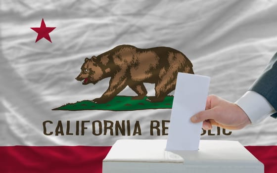 man putting ballot in a box during elections  in front of flag american state of california