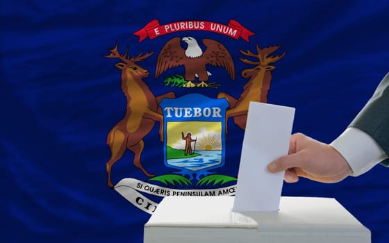 man putting ballot in a box during elections  in front of flag american state of michigan