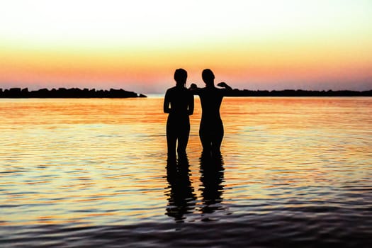 Silhouettes of two girls standing in the sea watching a tropical sunrise.