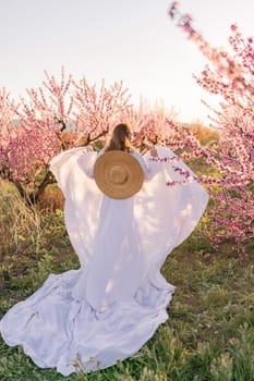 Woman blooming peach orchard. Against the backdrop of a picturesque peach orchard, a woman in a long white dress and hat enjoys a peaceful walk in the park, surrounded by the beauty of nature