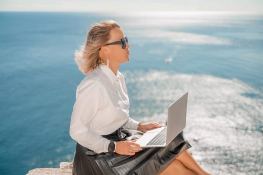 Business woman on nature in white shirt and black skirt. She works with an iPad in the open air with a beautiful view of the sea. The concept of remote work