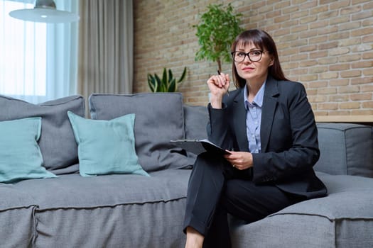 Portrait of confident mature female psychologist therapist counselor psychotherapist with clipboard sitting on couch, sofa in office. Mental health treatment therapy, assistance, support
