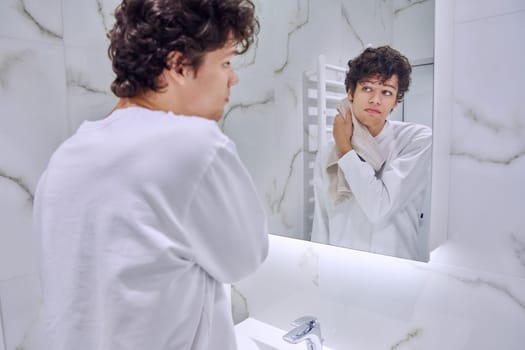Handsome young guy 18-20 years old in bathroom with towel looking at his reflection in mirror. Morning evening routine, hygiene, skin care facial cosmetics for young males