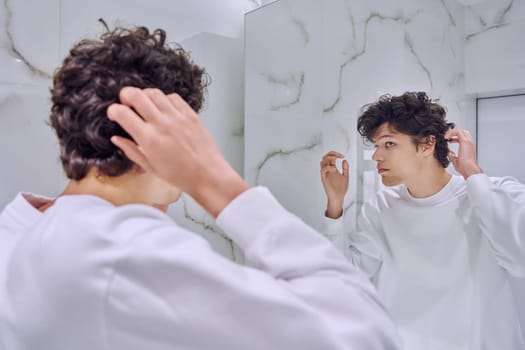 Young handsome male looking at himself in bathroom mirror, touching curly trendy hairstyle, admiring himself. Youth, fashion, beauty, hygiene, lifestyle, hair care, daily hair styling routine