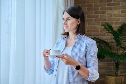 Young smiling woman with cup of morning coffee at home near window. Thoughtful daydreaming calm 30 year old female looking out the window, white curtain copy space