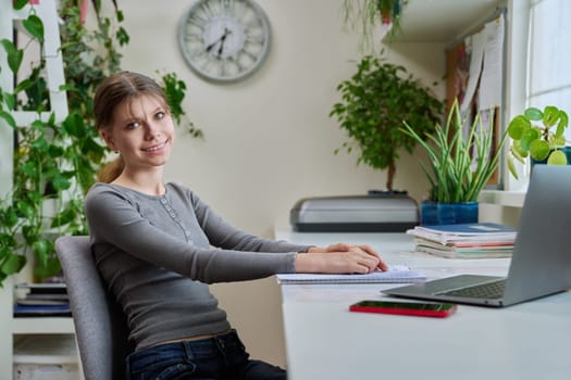 Portrait of teenage girl sitting at desk at home with laptop computer. Smiling 12, 13 year old young female student looking at camera. High school, adolescence, education, lifestyle concept