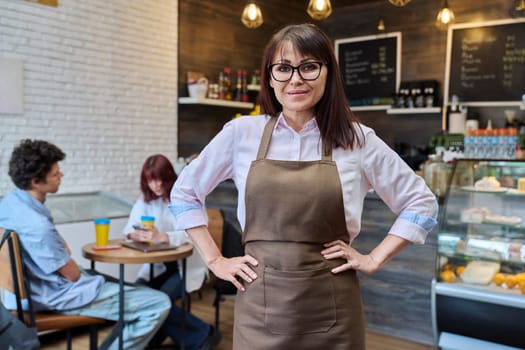 Portrait of confident smiling middle aged woman coffee shop owner, worker. Female in apron looking at camera inside cafe hall, customers drinking coffee, working barista. Small business, work, people