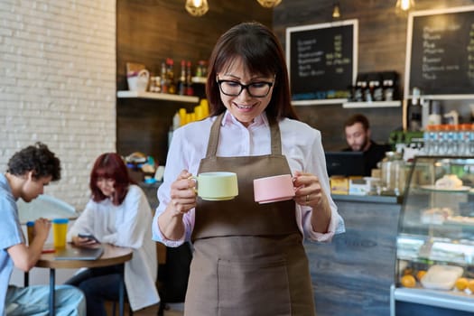 Portrait of smiling female coffee shop worker holding two cups of coffee in her hands. Woman in apron looking at camera inside cafe hall. Small business, food service occupation, staff, work concept