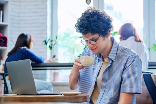 Handsome young curly-haired guy drinking cup of coffee in coffee shop. Young male college student in glasses enjoying cappuccino. Coffee culture, lifestyle, youth, people concept