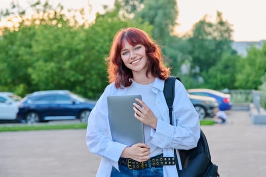 Outdoor portrait of young smiling female college student with backpack. Attractive girl with red hair holding laptop, looking at camera. 18-20 years students, youth, education, lifestyle concept