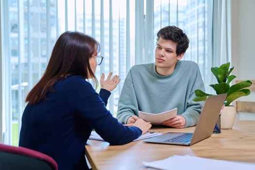 Middle aged woman teacher mentor talking helping testing male teenage student sitting in office in college building. Education, learning, contact teacher student concept