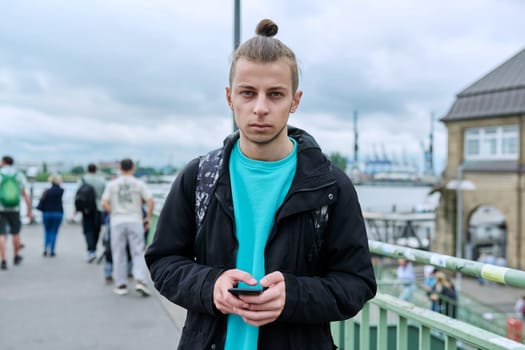 Portrait of handsome guy 19, 20 years old, modern city background. Young male with trendy beard hairstyle, with backpack, urban style. Youth, lifestyle, life concept