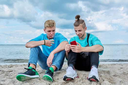 Young teenage male friends sitting on sand at beach, talking, laughing, looking in smartphone. Youth, summer vacation, communication, relaxing, leisure, friendship, fun, lifestyle holiday concept