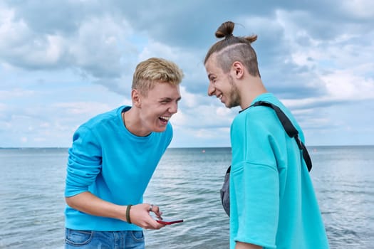 Two friends laughing cheerful young guys with smartphone, outdoor sea sky beach background. Men 18-20 years old age, youth, positive emotions leisure friendship communication, summer holidays concept