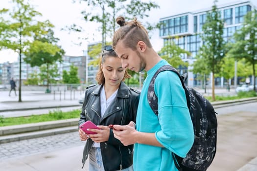 Teen friends guy and girl standing together holding smartphones looking at screen using mobile phones outdoor on city street. Internet digital technology applications for leisure study communication