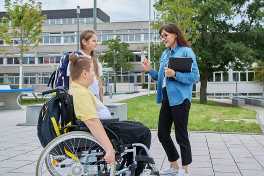 Female teacher with digital tablet and children classmates 10, 11 years old, boy in wheelchair and girl talking together outdoor, near school building. Education, disability, inclusiveness