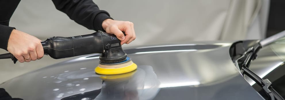 A mechanic polishes the surface of the hood of a gray car. Widescreen