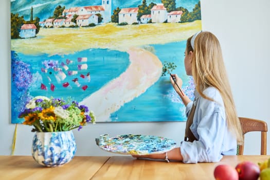 Young woman artist with acrylic paint palette with brushes painting picture on canvas on wall. Creativity, hobby, art therapy, leisure, paintings for sale, youth concept