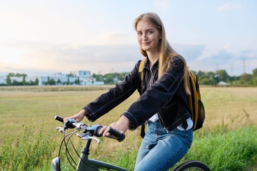Teenage female with backpack on bike looking at camera, outdoor nature road sunset sky background, copyspace. Activity, lifestyle, youth concept