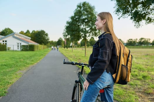 Portrait of teenage student girl with backpack on bicycle. Smiling teen female 16, 17 years old outdoors on road, copy space. High school, lifestyle, adolescence, youth concept