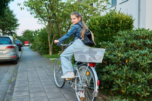 Back to school. Girl child 10, 11 years old with backpack on bicycle on street near house, posing looking at camera. Schoolgirl cycling to school, lifestyle, childhood concept