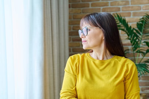 Middle-aged woman looking out the window, calm, thoughtful 40s female in yellow. Lifestyle, emotions, health middle age concept