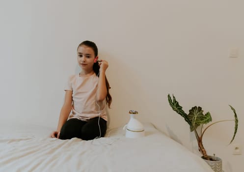 One beautiful Caucasian brunette girl with collected hair and in a pink T-shirt treats her ear with an infrared light device, sitting on her knees on a bed at home near a white wall, close-up side view.
