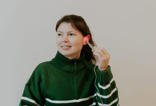 One beautiful Caucasian brunette girl with a smile on her face, gathered hair in a green sweater, treats her left ear with an infrared light device, sitting on a bed against a white wall, close-up side view.