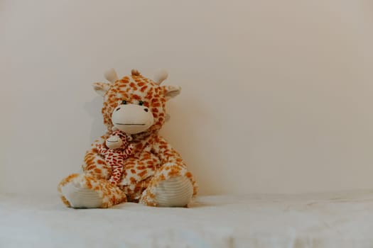 One beautiful soft toy giraffe sits on the left on the bed against the white wall with copy space on the right, close-up side view.