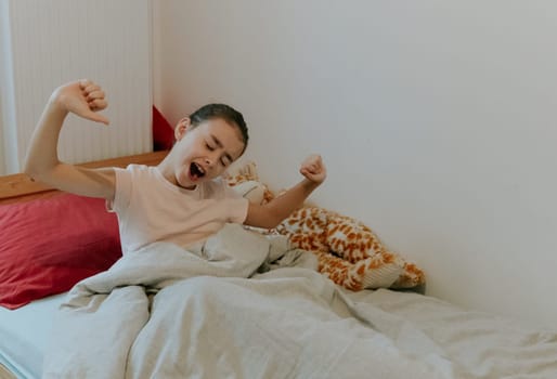 One beautiful little Caucasian brunette girl, waking up, yawns and sits up in bed with her eyes closed, stretches her arms out to the sides, close-up side view.