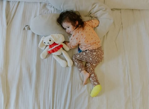 One beautiful little Caucasian girl with a cast on her leg lies on her stomach on the bed with a soft toy in the morning in the room, flat lay close-up.