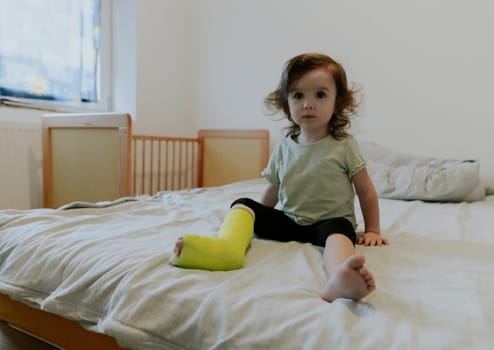 One beautiful little Caucasian baby girl with a green cast on her leg sits on the edge of the bed, watching a cartoon on TV in the morning in the room, side view close-up.