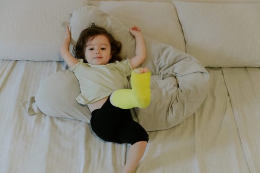 One beautiful little Caucasian girl baby with a cheerful emotion and a green cast on her leg lies on her back on the bed watching a cartoon on TV in the morning in the room, side view close-up.