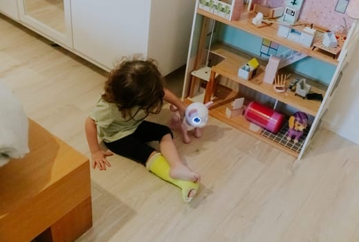 One Caucasian beautiful baby girl with a green cast on the floor with a sad emotion on her face plays with a toy while sitting on the wooden floor in the morning in the room, close-up top view.
