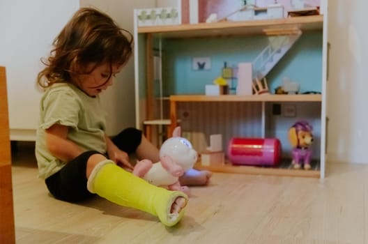 One Caucasian beautiful baby girl with a green cast on the floor with a sad emotion on her face plays with a toy while sitting on the wooden floor in the morning in the room, side view close-up.