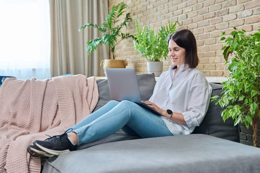 Young relaxed woman sitting on sofa at home in living room, using laptop for work, freelancing, learning, blogging, communicating, typing on keyboard