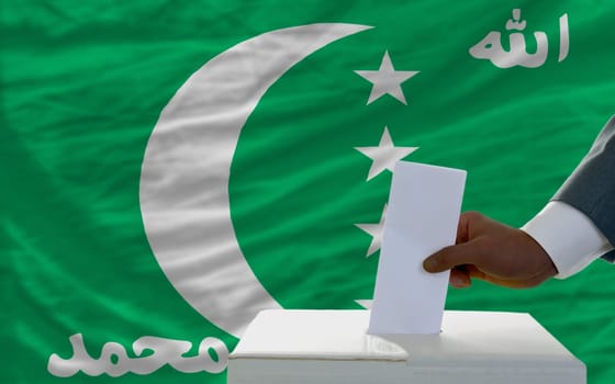 man putting ballot in a box during elections  in front of national flag of comoros