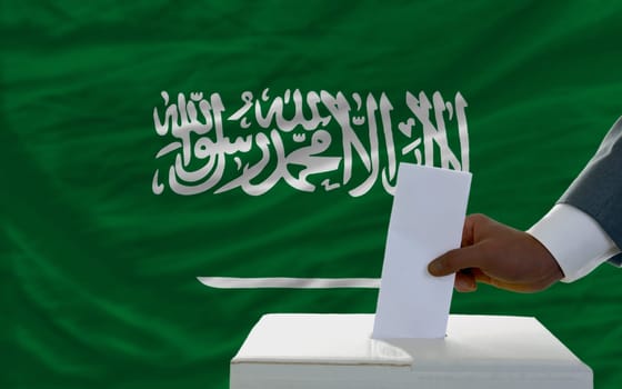 man putting ballot in a box during elections  in front of national flag of saudi arabia