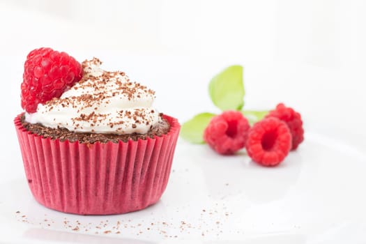 Chocolate cupcake topped with icing and a raspberry sprinkled with chocolate, raspberries in the distance.