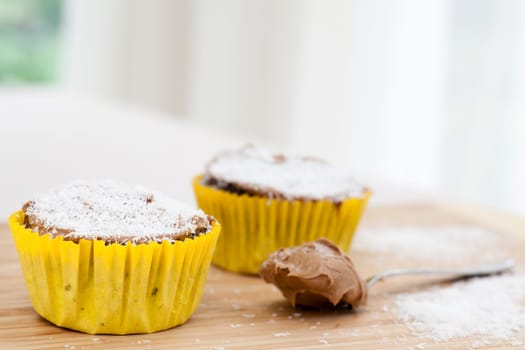 Two chocolate cupcakes sprinkled with coconut on cutting board with spoon and icing.