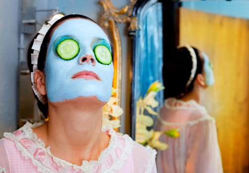 Funny housewife beauty green clay mask and cucumber at the mirror
