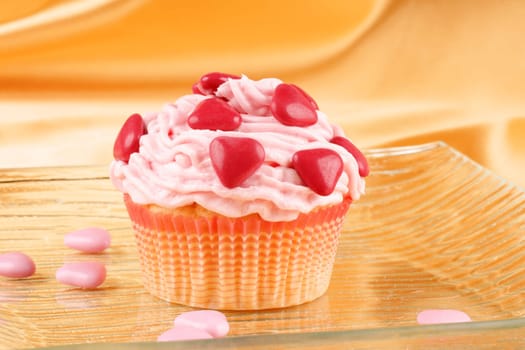 Fancy Valentine&#039 s Day cupcake decorated with small red sugar-coated chocolate hearts on a transparent glass dish, over a yellow background. Selective focus.