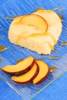 Heart shaped peach bavarian cream dessert with peach sauce and peach slices on a transparent glass plate over a blue background.