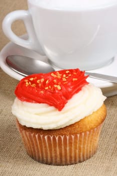 Fancy Valentine&#039 s day cupcake frosted with a red heart shape and a cappuccino over a natural jute background. Selective focus.