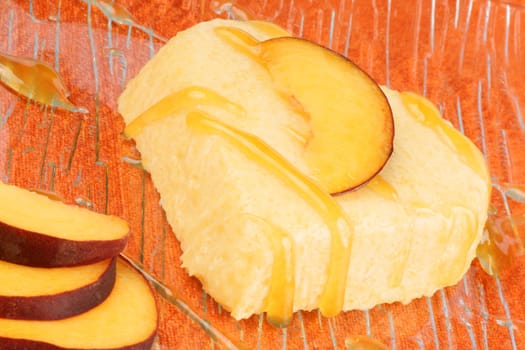 Heart shaped peach bavarian cream dessert with peach sauce and peach slices on a transparent glass plate over an orange background.