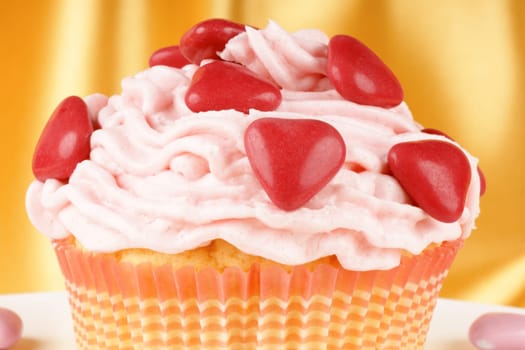 Fancy Valentine&#039 s Day cupcake decorated with small red sugar-coated chocolate hearts on a white plate, over a yellow background. Selective focus.